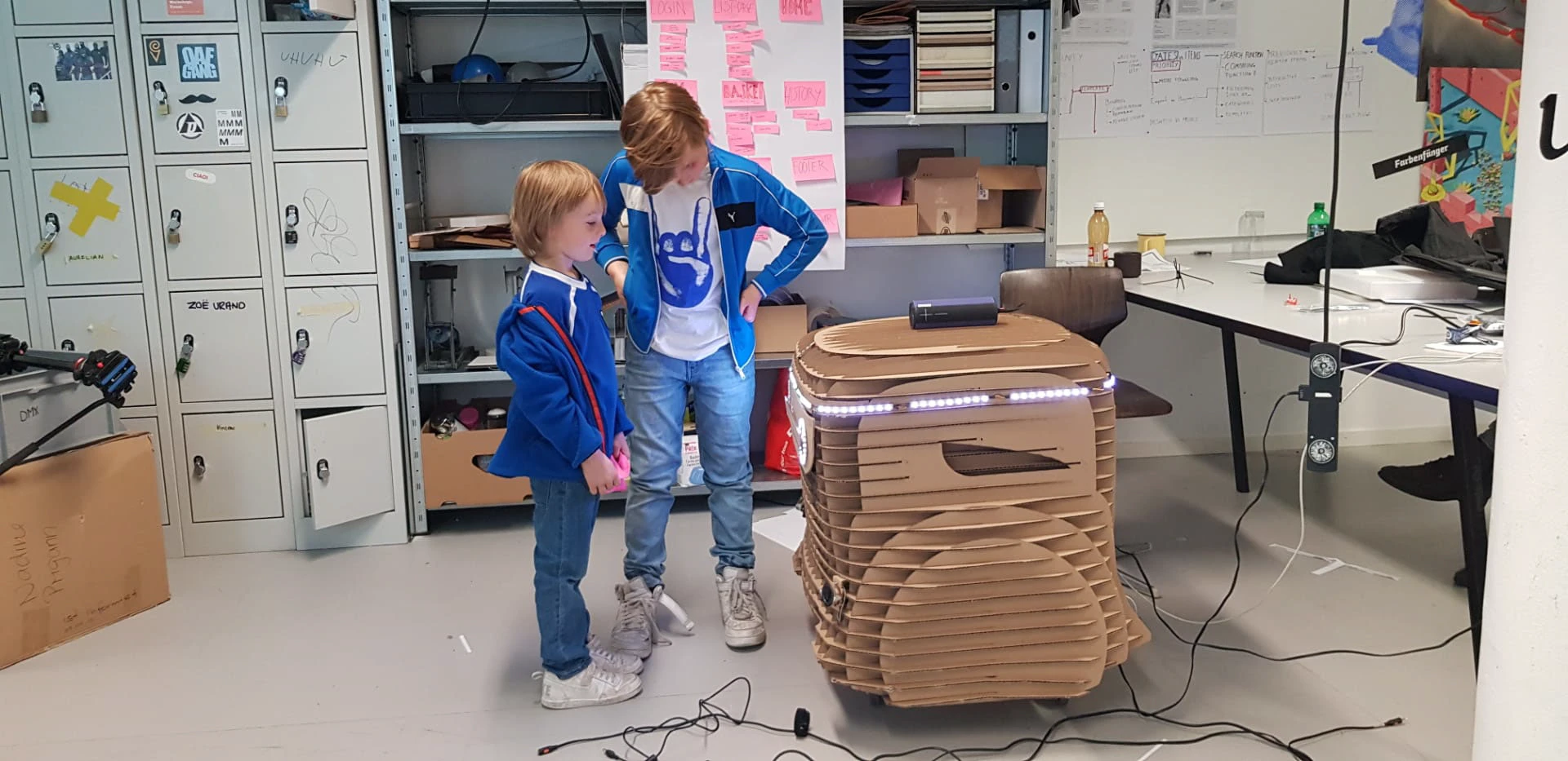 Two children standing next to the cardboard prototype in the studio, its LED strip lit, working out what it does.