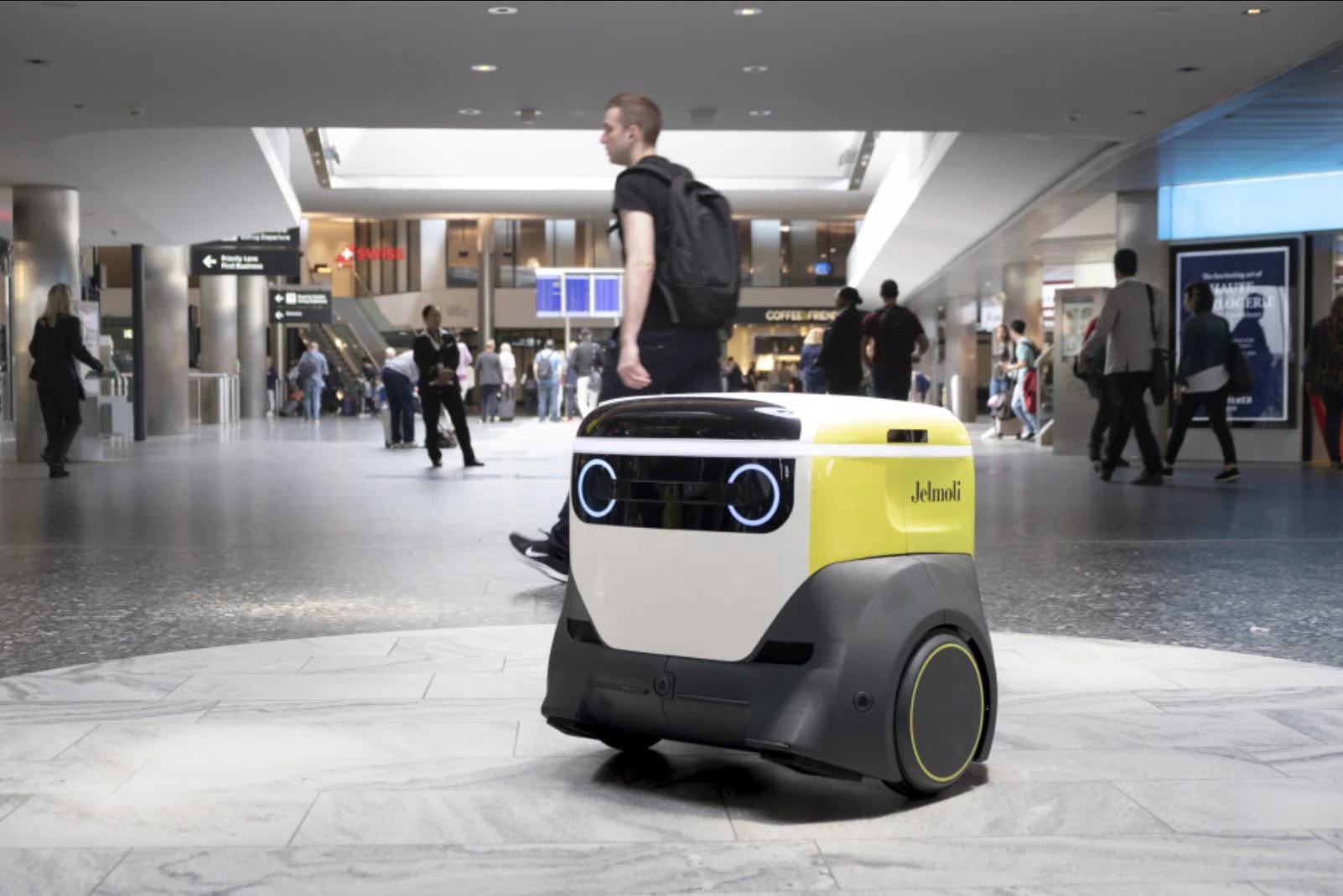 adero in the Zurich Airport concourse: a yellow, white and grey delivery robot with two lit ring eyes, travellers walking past it.