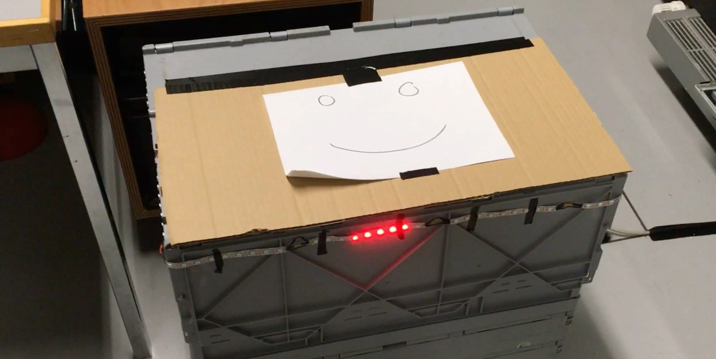 The first prototype: a grey folding crate with a cardboard lid, a hand-drawn smiley face taped to the top, and a red LED strip along the front edge.