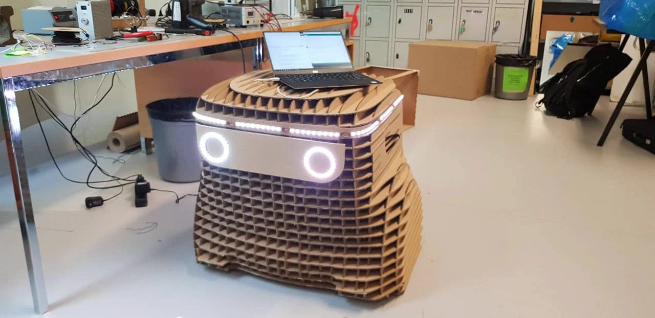 A full-size laser-cut cardboard prototype of adero in the workshop, two glowing white rings on the front and a lit bar above them, a laptop sitting on top driving it.