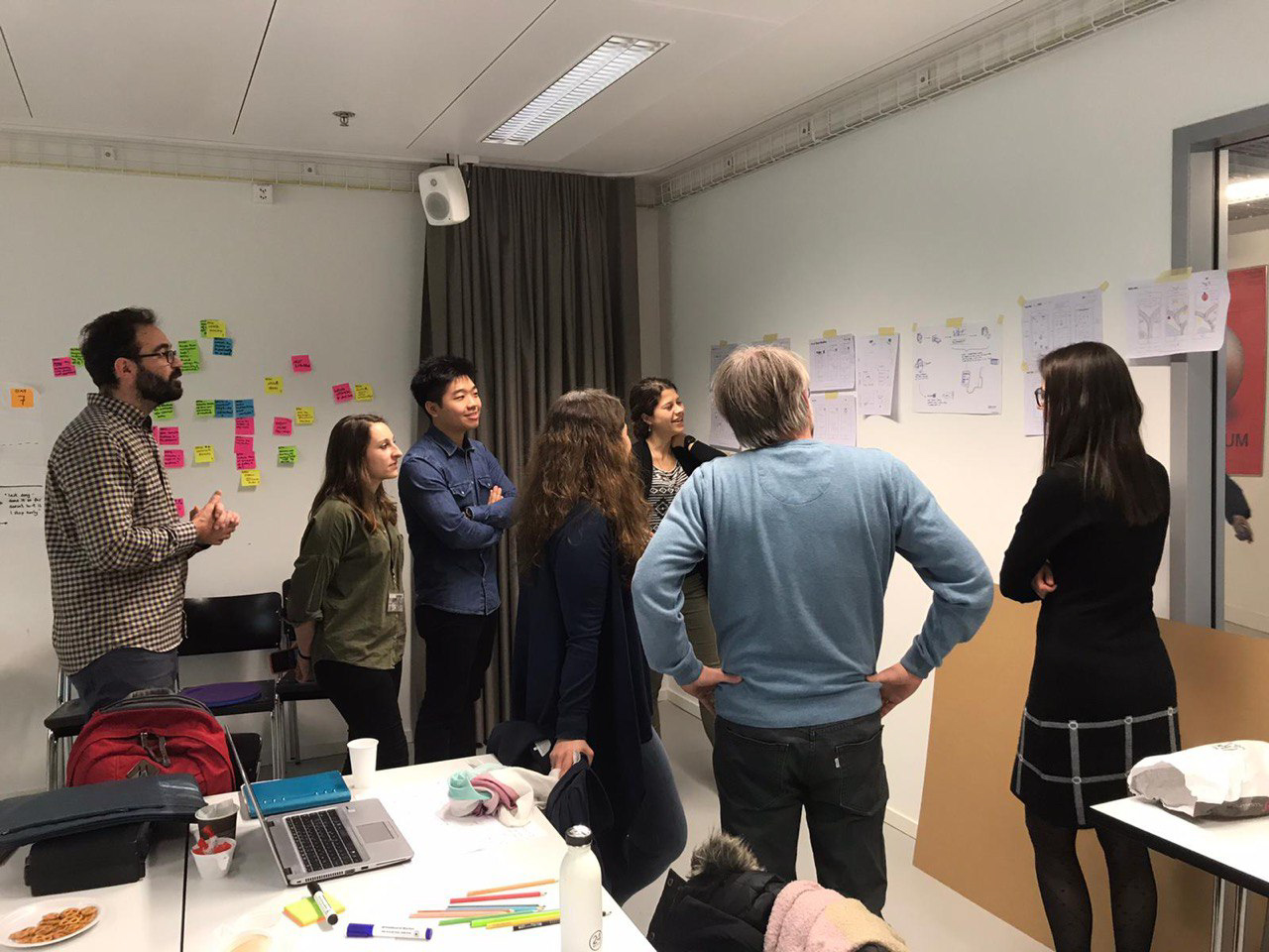 Designers and clinicians standing around a wall of sticky notes and printed sketches during a workshop.